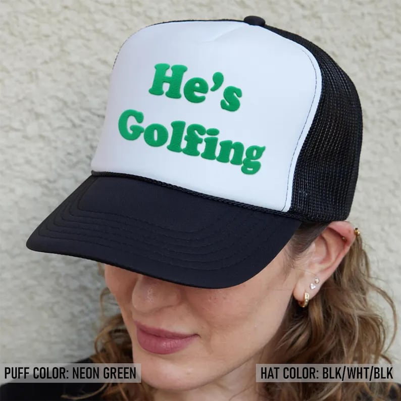 May include: A black and white trucker hat with a neon green puff print that says "He's Golfing".