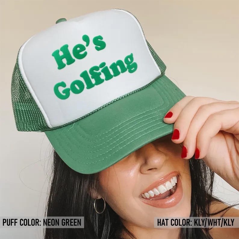 May include: A white and green trucker hat with the text "He's Golfing" in green letters. The hat has a mesh back and a green brim.