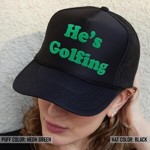 May include: Black trucker hat with a neon green puff print that says "He's Golfing".