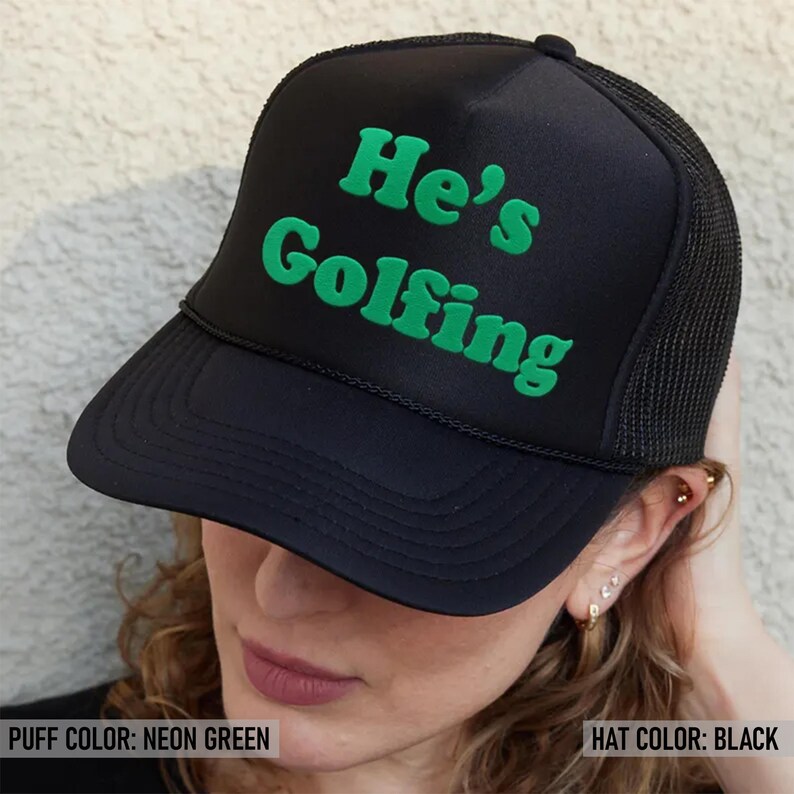 May include: Black trucker hat with a neon green puff print that says "He's Golfing".
