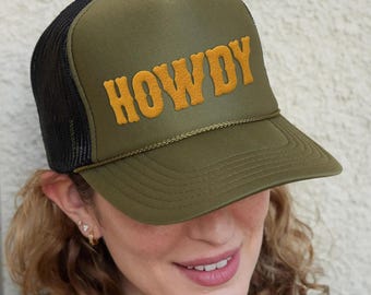 May include: An olive green trucker hat featuring a black mesh back and a structured front panel. The word HOWDY is embroidered in a raised, golden yellow puff font across the front. A thin, matching olive green cord trim sits above the brim.
