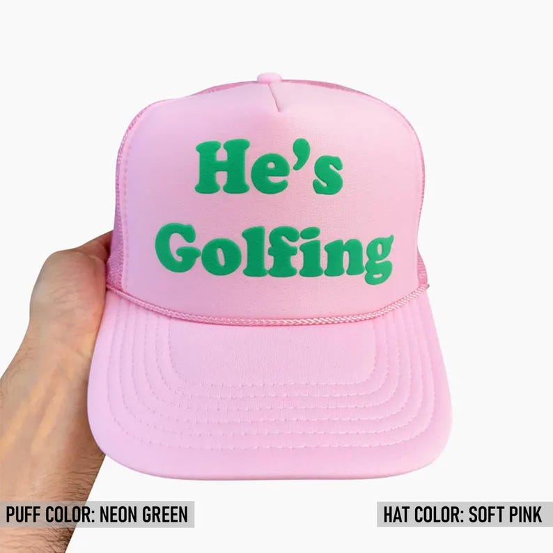 May include: A pink trucker hat with a neon green puff print that says "He's Golfing".