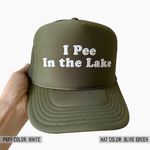 May include: An olive green trucker hat featuring a mesh back and a structured front panel. The front displays the white text "I Pee In the Lake" in a raised, puffy font. A thin, matching olive green cord runs horizontally across the base of the crown.