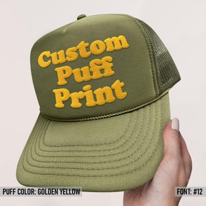 May include: An olive green trucker hat featuring a mesh back panel and a structured front panel. The front displays the text Custom Puff Print in raised, golden yellow lettering. A decorative cord runs along the base of the front panel.