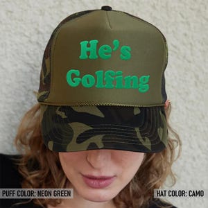May include: A green camouflage trucker hat with a black mesh back. The hat has neon green puff lettering that says "He's Golfing".
