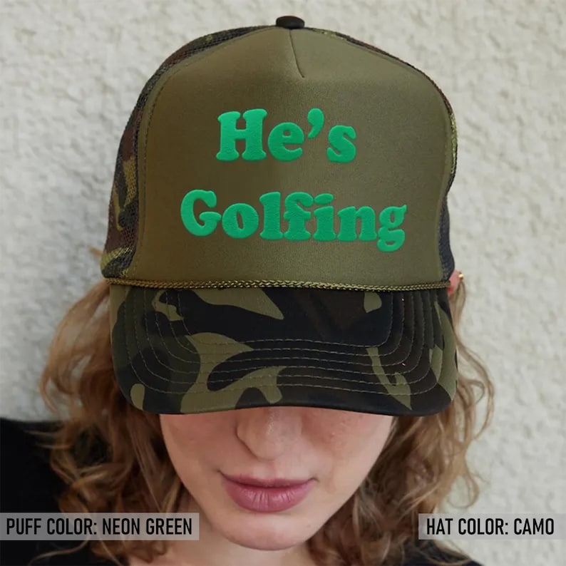 May include: A green camouflage trucker hat with a black mesh back. The hat has neon green puff lettering that says "He's Golfing".
