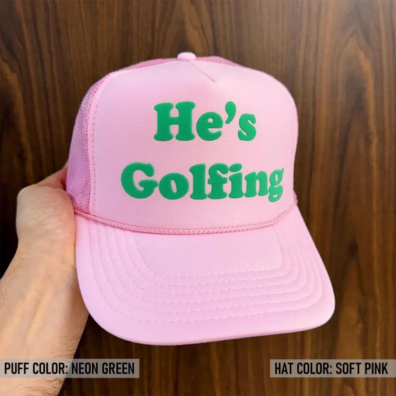 May include: A pink trucker hat with a green neon puff print that says "He's Golfing".
