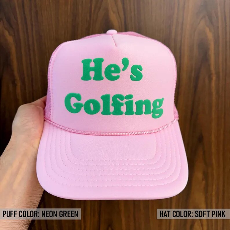 May include: A pink trucker hat with a neon green puff print that says "He's Golfing".