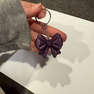 Pode incluir: Um porta-chaves roxo em forma de laço. O laço é feito de um material texturizado e é preso a uma corrente e um porta-chaves prateados. O porta-chaves é segurado contra uma superfície branca.