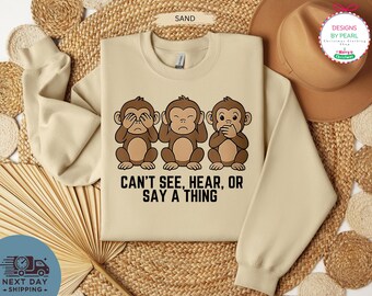 May include: A sand-colored sweatshirt featuring three cartoon monkeys in brown, each in a different pose: covering eyes, ears, and mouth. The text "CAN'T SEE, HEAR, OR SAY A THING" is printed below the monkeys.