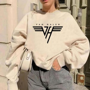 May include: Cream-colored sweatshirt featuring the band name "VAN HALEN" above a black and white logo. The sweatshirt is a relaxed fit and is paired with gray denim jeans. A white purse is visible.