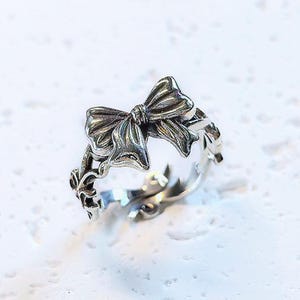 Può includere: Un anello argentato con un design a fiocco dettagliato. La fascia dell'anello presenta un motivo intricato di ispirazione floreale. Il fiocco ha dettagli strutturati e l'anello sembra essere in metallo.