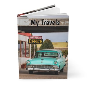 Peut inclure: Un livre à couverture rigide dont la couverture présente une voiture turquoise vintage garée devant un motel avec une enseigne indiquant Vacancy Office. Le texte My Travels est imprimé en noir en haut de la couverture, montrant un bâtiment.