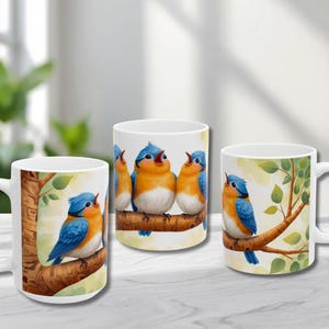 May include: Three white ceramic mugs featuring illustrations of bluebirds perched on tree branches. The birds have blue wings and heads, orange chests, and are singing. The mugs are set against a blurred background.