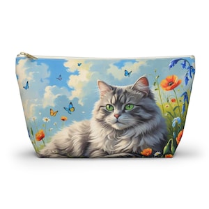 May include: A zippered cosmetic bag featuring a fluffy gray cat with green eyes, resting in a field of wildflowers and butterflies. The background depicts a blue sky with clouds. The bag has a light blue top and a gold zipper.