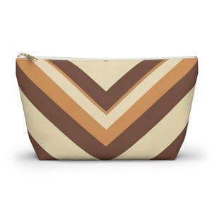 May include: A zippered pouch with a chevron pattern in shades of brown and beige. The pouch has a rectangular shape with a flat bottom and a gold-colored zipper. The design features alternating stripes of dark brown, light brown, and beige.