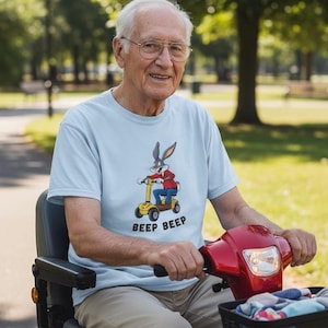 May include: A light blue t-shirt featuring Bugs Bunny riding a yellow scooter with the text "BEEP BEEP". The shirt is worn by a person sitting in a red mobility scooter. The person is wearing glasses and smiling.