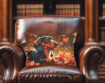 May include: A decorative pillow featuring a dachshund dog lying in autumn leaves. The pillow has a black, brown, and orange color scheme. The pillow is placed on a brown leather armchair in front of a bookshelf.