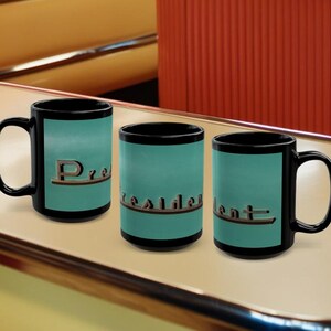 May include: Three black coffee mugs with a teal blue background and the word "President" in gold lettering. The mugs are arranged in a row on a light brown surface.