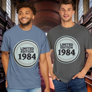 May include: Two people wearing t-shirts with a circular design that reads "LIMITED EDITION 1984". One shirt is blue, the other is grey. The background shows a library with bookshelves.