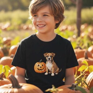 May include: A black t-shirt featuring a graphic print of a brown and white puppy sitting next to an orange jack-o'-lantern pumpkin. The design is centered on the chest of the garment, showcasing a realistic illustration of the dog and pumpkin.
