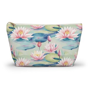 May include: A zippered pouch featuring a watercolor pattern of pink and white water lilies, lily pads, and buds on a light green background. The pouch has a gold-colored zipper and is designed for carrying cosmetics or other small items.