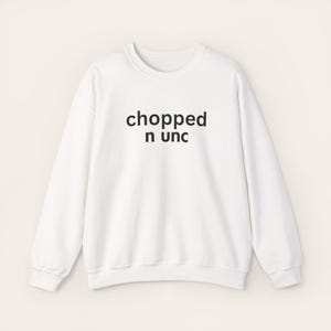 May include: A white sweatshirt featuring the black text "chopped n unc" in a centered, stacked design. The long-sleeved top has a round neck, ribbed cuffs and hem. The background is a plain, light color.