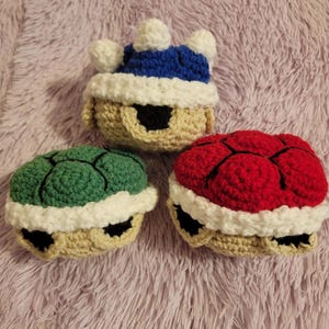 May include: Three crocheted turtle plushies with different colored shells. The first turtle has a blue shell with a white crown, the second turtle has a green shell, and the third turtle has a red shell.