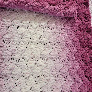 May include: A handmade, plush blanket in shades of pink and white. The blanket features a textured, shell-like stitch pattern. The colors transition from a deep magenta at the top to a soft white at the bottom, creating a gradient effect. The blanket is folded over at the top.