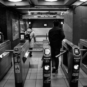 May include: Black and white image of a subway station entrance with turnstiles. The turnstiles have digital displays and the text "tapp AND RIDE HERE". Two people are walking through the turnstiles. The sign above reads "To PATH Trains".