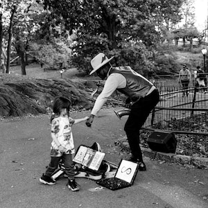 Può includere: Fotografia in bianco e nero di un musicista di strada che suona il sassofono in un parco. Un bambino si avvicina al musicista. Un computer portatile con un simbolo e attrezzature musicali sono a terra.