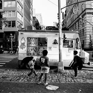 Puede incluir: Escena callejera en blanco y negro con un camión de helados y niños. El camión muestra gráficos de conos de helado y varios productos. Se ven edificios y un paso de cebra en el fondo.