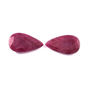 May include: Two pear-shaped, faceted rubies with a deep magenta-red hue. Each gemstone features a smooth, polished surface with visible internal inclusions and a pointed end, displaying a consistent colour saturation across both individual stones.