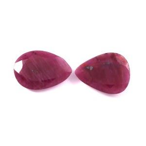 May include: Two pear-shaped, faceted rubies with a deep magenta-red hue. Each gemstone displays a smooth, polished surface with visible internal inclusions and a distinct, multi-faceted cut that reflects light across the top surfaces.