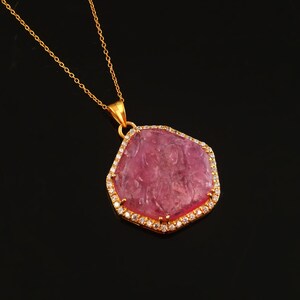 May include: A gold necklace featuring a large, irregularly shaped pink pendant. The pendant is edged with small, clear stones and has a textured surface. The necklace is set against a black background.