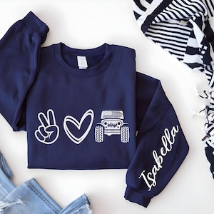 May include: Navy blue sweatshirt with a white graphic of a peace sign, heart, and a jeep with an American flag. The right sleeve has the name "Isabella" in white script. The sweatshirt is paired with blue jeans.