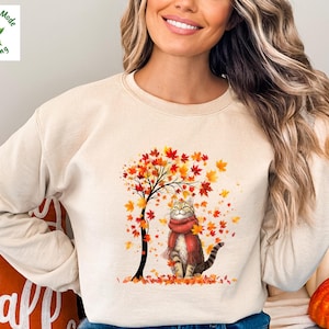 May include: Beige sweatshirt featuring a cat wearing a red scarf, sitting under a tree with autumn leaves. The graphic includes a tree with red and orange leaves, and the text "Magnolia Mode Clothing" in a green box.