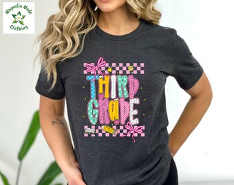 Op de afbeelding: Donkergrijs t-shirt met een kleurrijke "Third Grade" afbeelding. Het ontwerp bevat een roze strik, een potlood en een dambordpatroon. Het shirt is van Magnolia Mode Clothing.