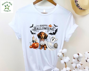 May include: A white short-sleeve t-shirt featuring a graphic of a dachshund wearing a white ghost costume. The design includes the text "HAPPY HALLOWEENIE" with illustrations of pumpkins, lit candles, bats, a ghost, and a tombstone in orange, black, and white.