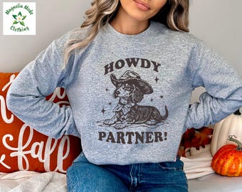 May include: A light gray sweatshirt featuring a dachshund wearing a cowboy hat and bow tie, with the words "HOWDY PARTNER!" printed below. The sweatshirt has long sleeves and a crew neck. The Magnolia Mode Clothing logo is in the upper left corner.