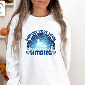May include: A white long-sleeve sweatshirt featuring a blue graphic print of a witch flying on a broomstick against a moonlit forest background. The text reads SUPPORT YOUR LOCAL WITCHES in a bold blue font, with small pumpkin icons on the sides.