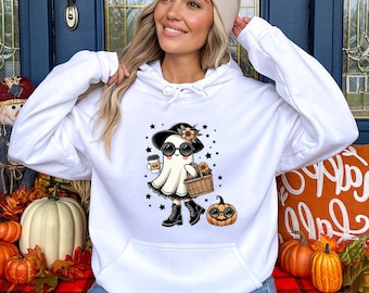 May include: White hooded sweatshirt featuring a cartoon ghost character wearing a hat, sunglasses, and boots, holding a coffee cup and a basket. A small pumpkin is next to the ghost. Black stars are scattered around the ghost.