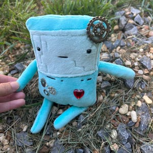 May include: A turquoise plush toy robot with a light blue face and arms. The robot has a red heart-shaped gem on its chest and a bronze gear on its head. Additional gears and gems are on the robot's body.