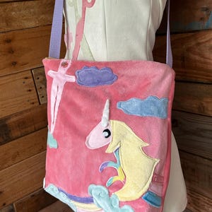 May include: Pink plush shoulder bag with a unicorn design. The bag features a unicorn with a yellow mane, a rainbow, and blue and purple clouds. The bag has a light purple strap and is made of soft, plush material. The bag is a whimsical accessory.