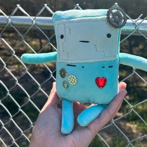May include: A light blue plush toy with a square head, small arms, and legs. It has a red heart-shaped gem on its chest and decorative gears. The toy is held in a person's hand.