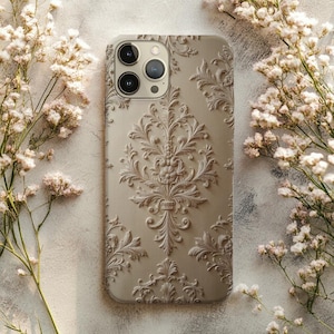 May include: A beige phone case with a detailed, raised floral pattern. The case has a matte finish and a camera cutout. The design is reminiscent of classic wallpaper. The case is surrounded by delicate white flowers.