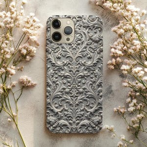 May include: A gray phone case with an ornate, raised floral pattern. The case has a matte finish and features a camera cutout. The background includes white flowers and a neutral surface.