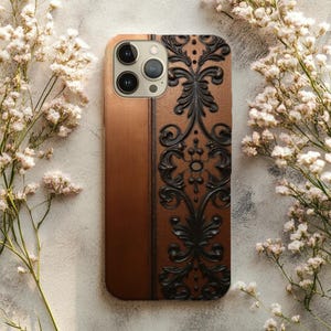 May include: A copper-colored phone case with a black, ornate, embossed design. The case has a vertical, textured panel on the left side and a decorative, symmetrical pattern on the right. The phone case is surrounded by white flowers.