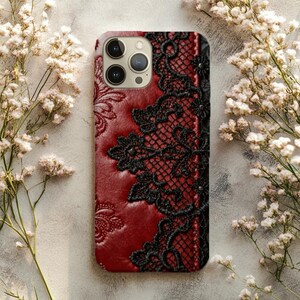 May include: A red phone case with a black lace design. The case has a textured, leather-like appearance and features a floral pattern on the left side. The black lace is intricately detailed, adding a touch of elegance to the phone accessory.