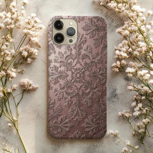 Puede incluir: Funda de teléfono de color malva con un patrón floral en relieve detallado. La funda tiene un acabado brillante y un recorte para la cámara. El diseño simétrico y ornamentado presenta hojas y elementos florales. Rodeado de flores blancas.